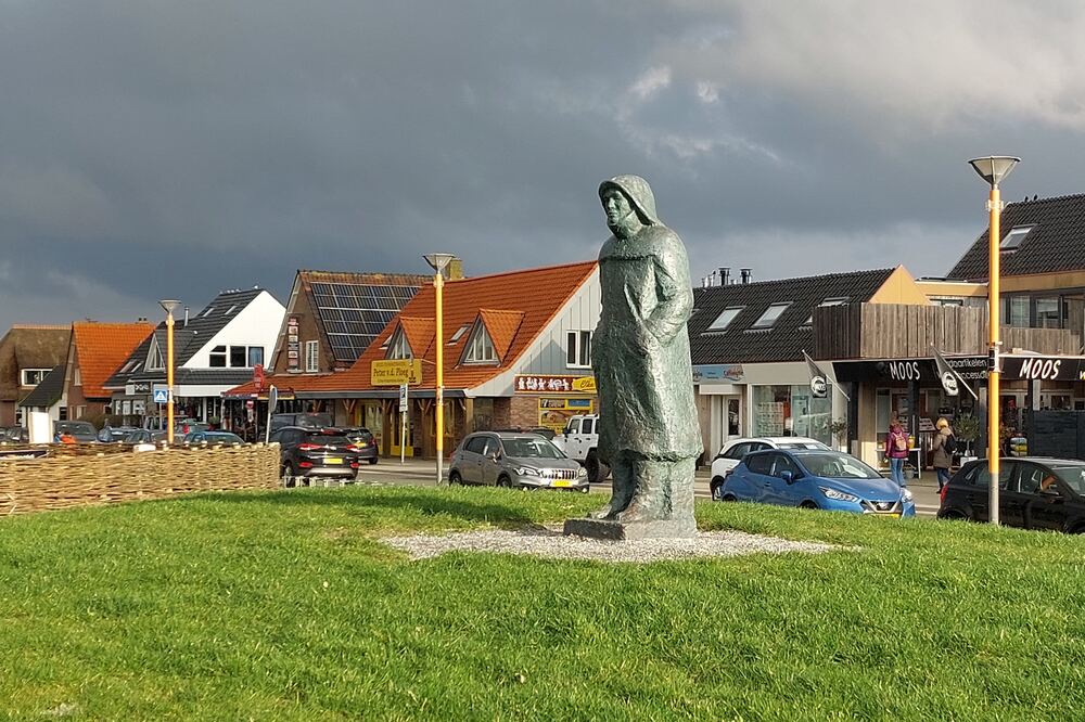 Bronzen beeld van een visser op een grasveld in het centrum van Callantsoog, met winkels en terrassen op de achtergrond
